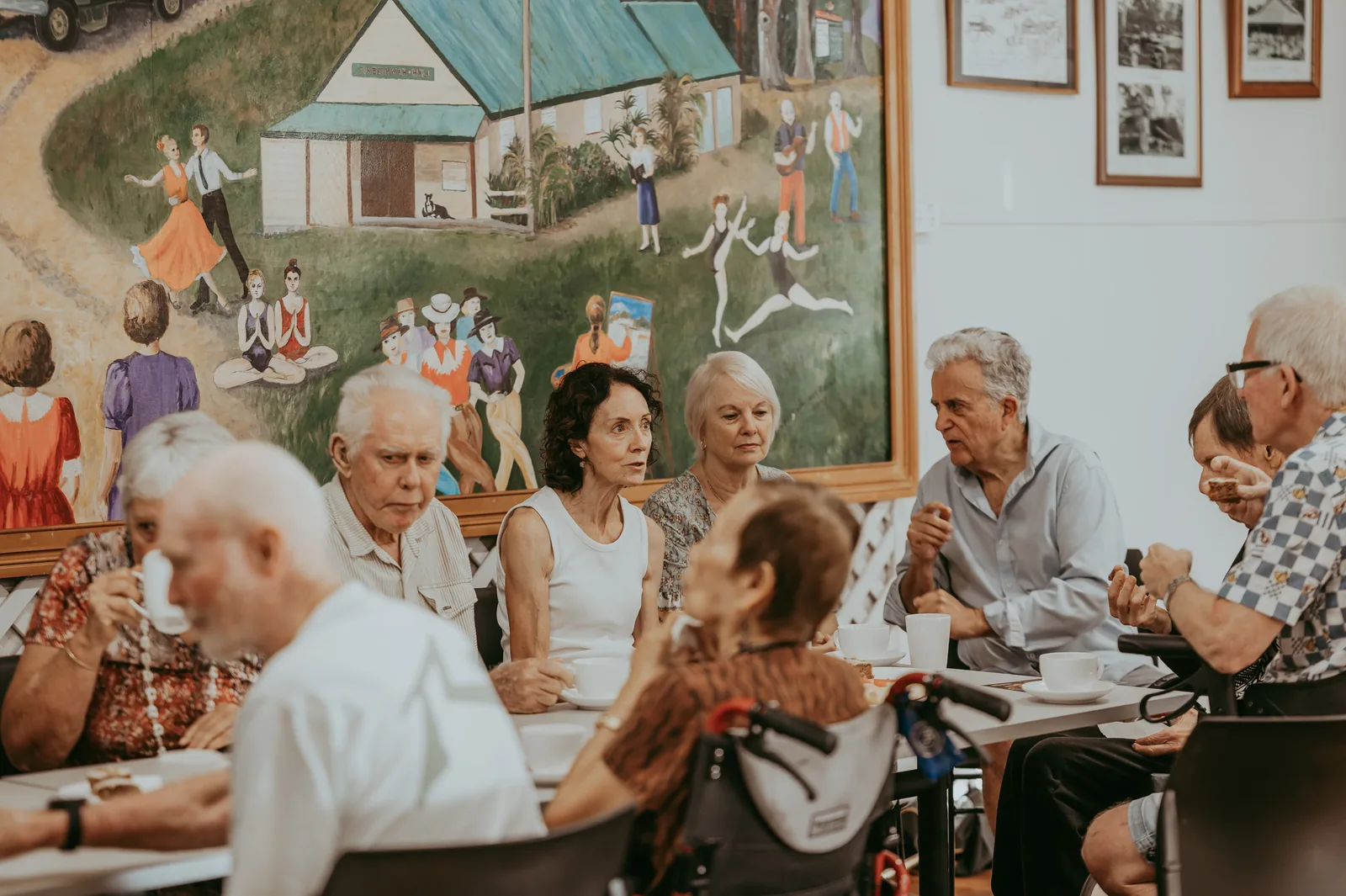 Members of The Sing Thing gathered around tables for afternoon tea, with a community-hall mural of dancing figures behind them.